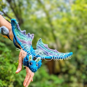 May include: A crocheted dragon toy featuring a blue head with small green horns, dark eyes, and large wings with a gradient of blue, purple, and green yarn. The text Lisa Bee Stitchin is overlaid in white script across the centre of the dragon.