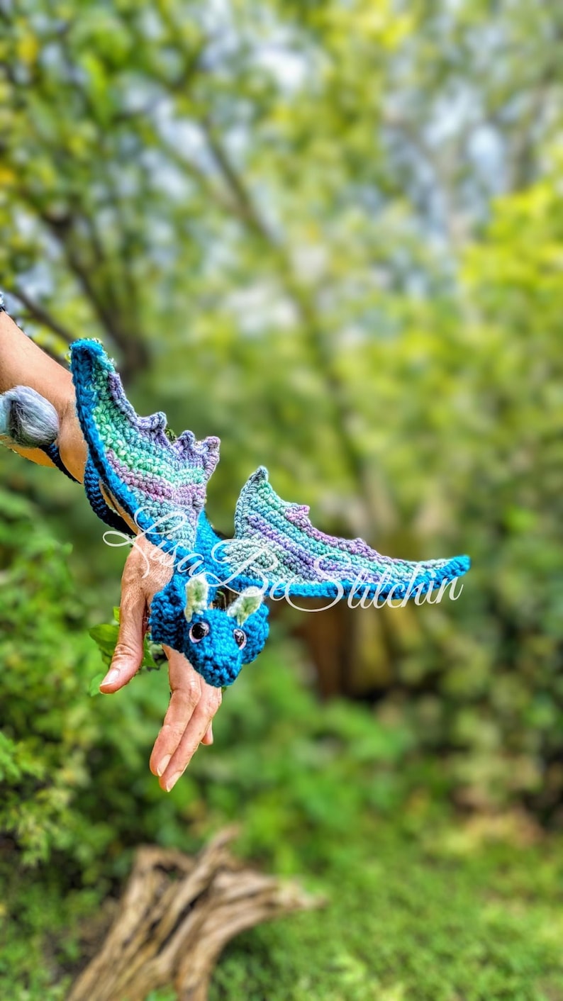 May include: A crocheted dragon toy featuring a blue head with small green horns, dark eyes, and large wings with a gradient of blue, purple, and green yarn. The text Lisa Bee Stitchin is overlaid in white script across the centre of the dragon.