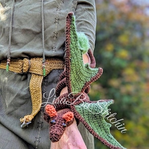 May include: A crocheted dragon accessory in dark brown with light green wings and orange ears. The dragon is designed to wrap around an arm, featuring a textured yarn construction, dark bead eyes, and a long tail that coils along the forearm.