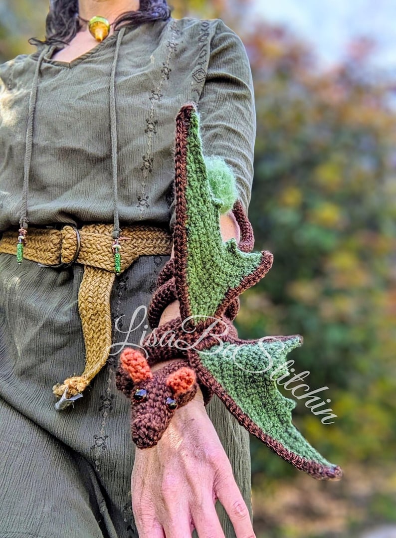 May include: A crocheted dragon accessory in dark brown with light green wings and orange ears. The dragon is designed to wrap around an arm, featuring a textured yarn construction, dark bead eyes, and a long tail that coils along the forearm.