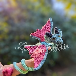 May include: A crocheted dragon toy featuring a light blue body with pink inner wings and a pink tail tip. The dragon has black horns, yellow eyes, and a long, coiled tail. The text "Lisa Ba Stitchin" is overlaid across the centre of the dragon.