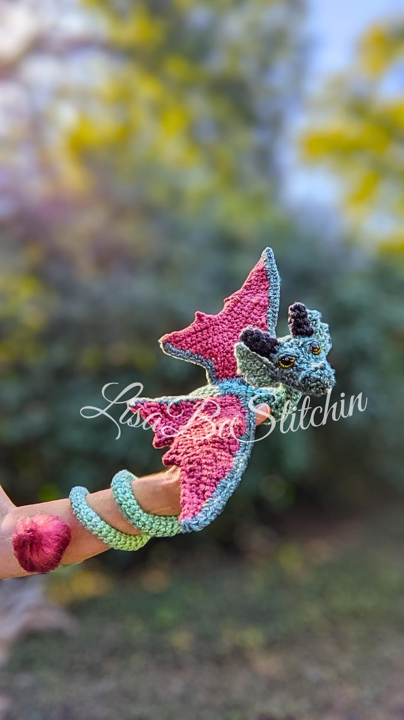May include: A crocheted dragon toy featuring a light blue body with pink inner wings and a pink tail tip. The dragon has black horns, yellow eyes, and a long, coiled tail. The text "Lisa Ba Stitchin" is overlaid across the centre of the dragon.