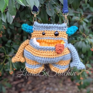 Pode incluir: Bolsa de monstro de crochê feita à mão com uma alça azul. A bolsa apresenta um corpo listrado azul, amarelo e cinza, chifres azuis e uma língua vermelha. A bolsa tem fecho de zíper e ferragens douradas. O texto "Lisa Be Stitchin" é visível.