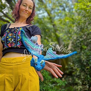 May include: A small, handcrafted crochet dragon toy featuring a vibrant blue body, light green horns, and wings with a gradient of blue, purple, and green yarn. The dragon has dark safety eyes and is displayed in a horizontal, flying position.