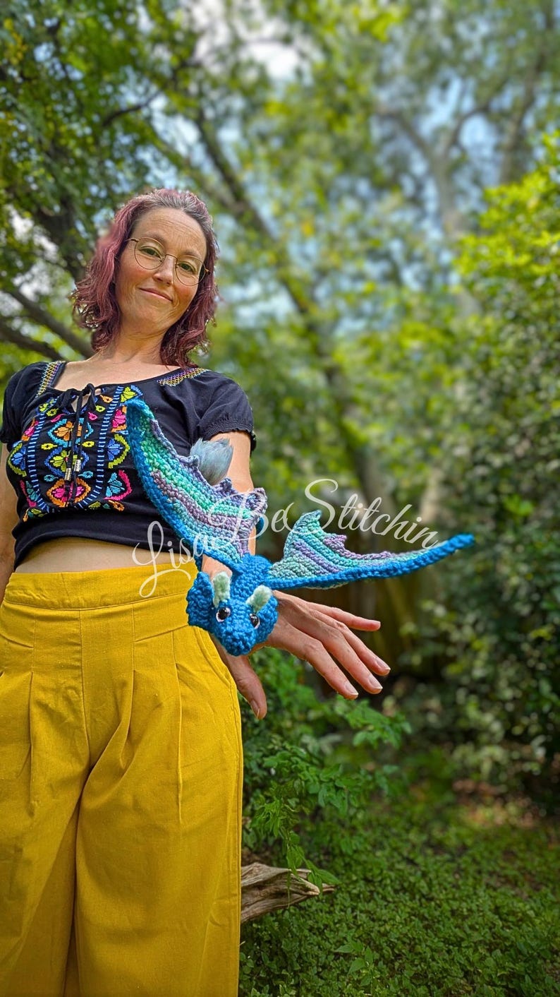 May include: A small, handcrafted crochet dragon toy featuring a vibrant blue body, light green horns, and wings with a gradient of blue, purple, and green yarn. The dragon has dark safety eyes and is displayed in a horizontal, flying position.