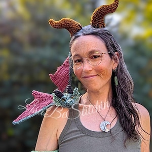 May include: A crochet dragon accessory featuring a blue and green head with wings resting on the shoulder, a long tail wrapped around the upper arm with a pink tuft, and two brown textured horns positioned above the head. The text Lisa Be Stitchin is visible.