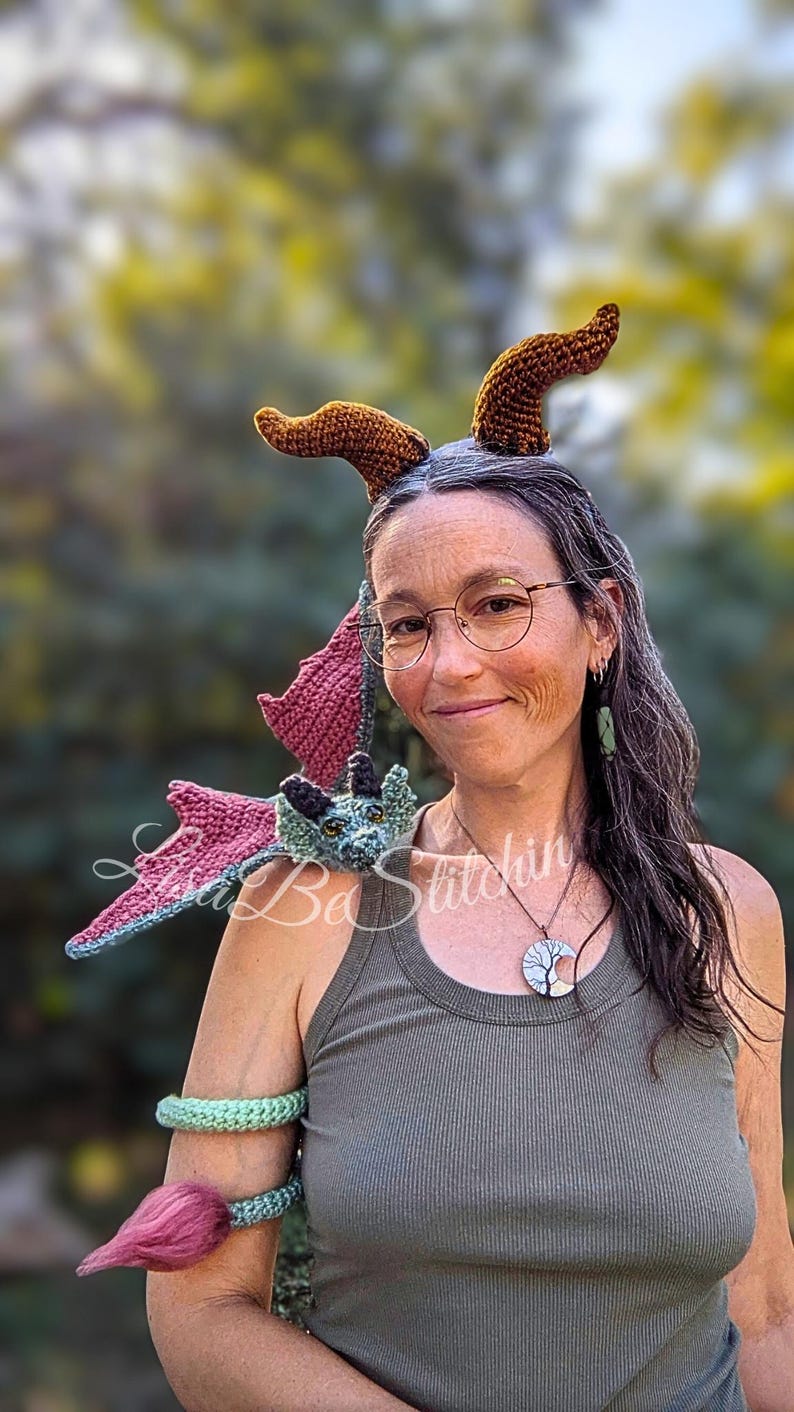 May include: A crochet dragon accessory featuring a blue and green head with wings resting on the shoulder, a long tail wrapped around the upper arm with a pink tuft, and two brown textured horns positioned above the head. The text Lisa Be Stitchin is visible.