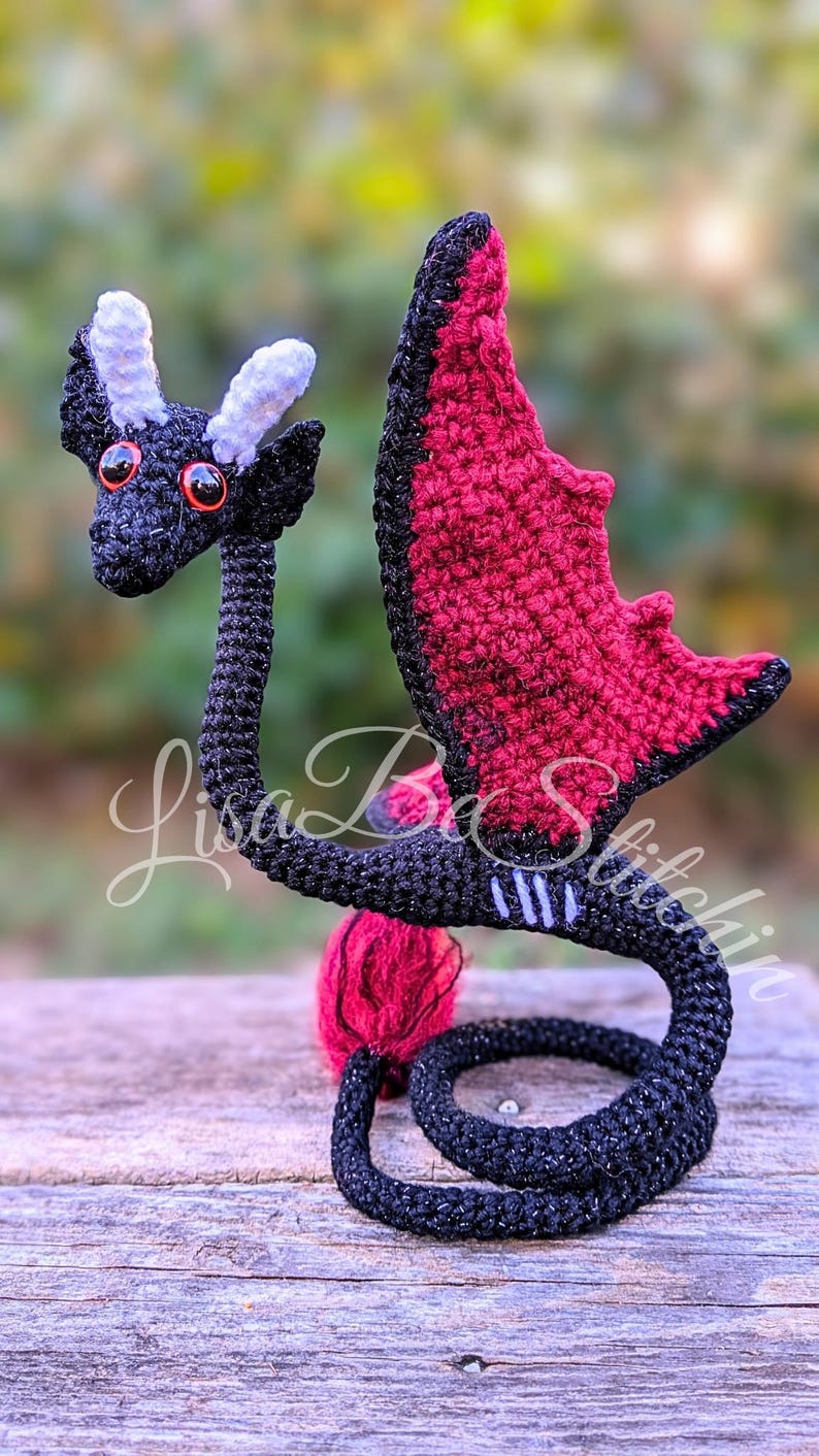 May include: A black and dark red crocheted dragon figurine with two light purple horns and bright orange eyes. The dragon has a long neck, a curled tail, and wings with a dark red interior. The text Lisa B Stitchin is overlaid on the image.