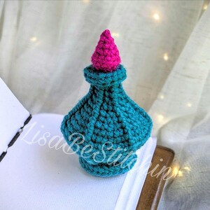 May include: A teal and pink crochet bottle with a rounded base and a pointed top. The bottle is placed on a white surface next to an open book. The text "LisaBestitchin" is visible on the image.