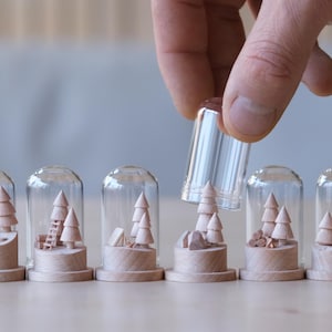 Peut inclure: Une main soulève un dôme en verre transparent d'une base en bois. À l'intérieur des dômes, des arbres en bois miniatures et des scènes de camping. Les bases sont sur une surface marron clair. Thème : forêt ou camping.