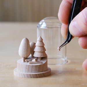 Peut inclure: Une scène miniature en bois représentant un paysage avec des arbres et une petite bûche, posée sur une base circulaire. Une pince à épiler tient une petite pièce en bois, peut-être pour la scène. Un dôme en verre transparent est en arrière-plan.
