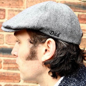 May include: A gray tweed flat cap with a black embroidered "Dad" on the side. The cap is worn by a person with dark hair.
