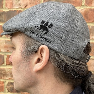 May include: A gray tweed flat cap with a black embroidered design that says "DAD The Fisherman" with a fish and fishing hook.