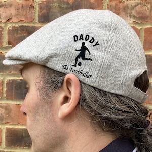 May include: A gray flat cap with a black silhouette of a person playing soccer and the text "Daddy The Footballer" on the front.