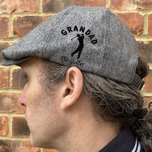 May include: Gray tweed flat cap with a black embroidered golfer graphic and the text "Grandad The Golfer".