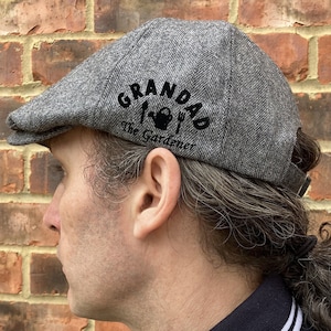 May include: A gray tweed flat cap with a black embroidered design that says "Grandad The Gardener" with a watering can, fork, and trowel.