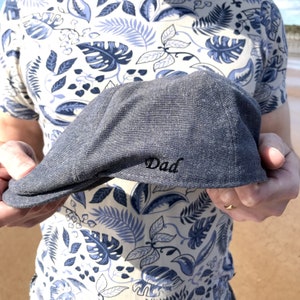 Könnte beinhalten: Graue Flatcap mit dem Wort "Dad" in Schwarz gestickt. Die Kappe wird vor einer Person gehalten, die ein Hemd mit blauem und weißem Blumenmuster trägt. Die Kappe ist aus einem strukturierten Stoff gefertigt.