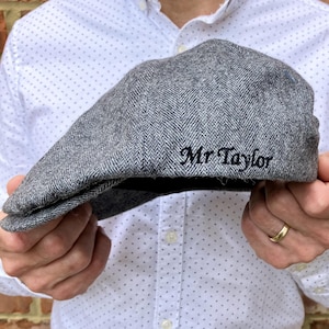 May include: A gray herringbone tweed newsboy cap with the embroidered name "Mr Taylor" in black script. The cap is held up in front of a white shirt with a small polka dot pattern.