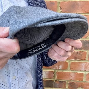 May include: A gray tweed flat cap with a black band. The band has the text "We love you Dad from Jack and Ellie x" embroidered in white.