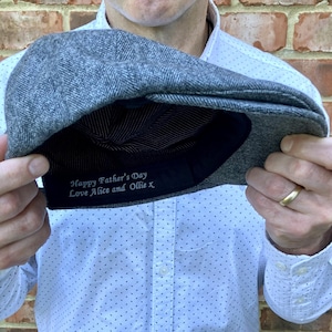 Könnte beinhalten: Graue Tweed-Newsboy-Cap mit dunklem Innenfutter. Auf der Innenseite der Kappe steht der Text "Happy Father's Day Love Alice and Ollie X". Die Kappe wird von einer Person gehalten, die ein weißes Hemd trägt.