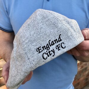 May include: A gray tweed flat cap with black embroidery that reads "England City FC".