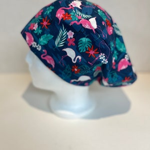 May include: A navy blue scrub cap featuring a tropical pattern with pink flamingos, white birds, and colorful flowers. The design includes green leaves and is set against a dark blue background. The cap is displayed on a white mannequin head.