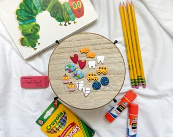 May include: A collection of school supplies and children's items, including a copy of "The Very Hungry Caterpillar" book, Crayola crayons, pencils, erasers, glue sticks, and decorative embellishments. The items are arranged on a white surface.