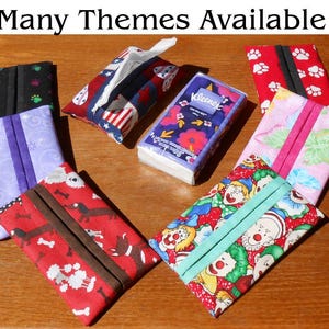 May include: A collection of rectangular fabric tissue holders in various patterns including red with dogs, purple with swirls, black with paw prints, red with white paw prints, and a clown print, surrounding a single pack of Kleenex On the Go tissues.