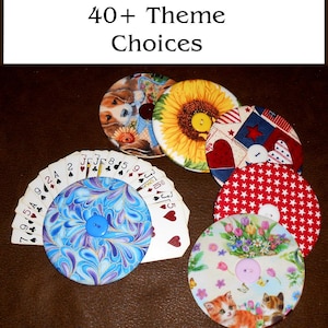 May include: A collection of round fabric coasters with various designs, including a sunflower, patriotic stars and hearts, kittens, and a blue and purple swirl pattern. A deck of playing cards is fanned out behind one coaster. The text "40+ Theme Choices" is at the top.