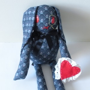 Może przedstawiać: Niebieski królik z tkaniny z czarną muszką i czerwonymi haftowanymi oczami trzyma białe koronkowe serce z czerwonym filcowym środkiem.