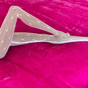 Könnte beinhalten: Weiße Netzstrümpfe mit Polka-Dot-Muster. Die Strümpfe werden auf einer leuchtend rosa Oberfläche präsentiert, was einen auffälligen Kontrast erzeugt. Das Netzdesign verleiht dem Kleidungsstück eine besondere Textur und visuelles Interesse.