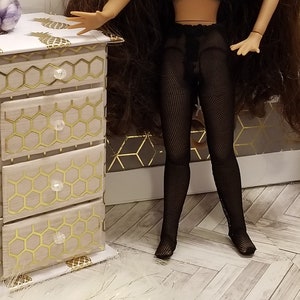 May include: A miniature dollhouse dresser with a white and gold pineapple design on top and gold honeycomb pattern on the drawers. The doll is wearing black fishnet stockings. The room has a white and gray color scheme.