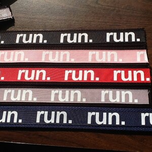 K&ouml;nnte beinhalten: Eine Sammlung von Stoffb&auml;ndern in verschiedenen Farben, auf denen jeweils das Wort "run." in wei&szlig;en Blockbuchstaben steht. Zu den Farben geh&ouml;ren Schwarz, Rosa, Rot, Grau und Marineblau. Die B&auml;nder sind nebeneinander angeordnet.