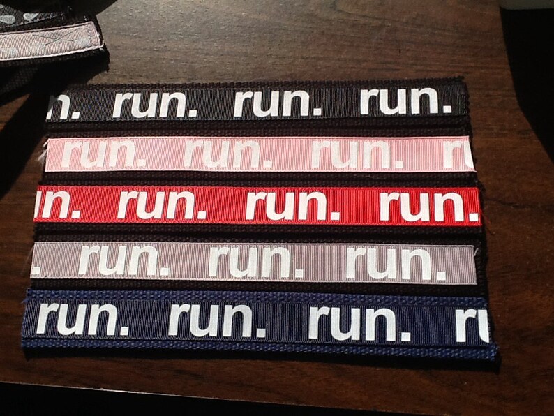 K&ouml;nnte beinhalten: Eine Sammlung von Stoffb&auml;ndern in verschiedenen Farben, auf denen jeweils das Wort "run." in wei&szlig;en Blockbuchstaben steht. Zu den Farben geh&ouml;ren Schwarz, Rosa, Rot, Grau und Marineblau. Die B&auml;nder sind nebeneinander angeordnet.