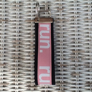 K&ouml;nnte beinhalten: Ein schwarzer und rosa Schl&uuml;sselanh&auml;nger mit dem Wort "run." in wei&szlig;en Buchstaben. Der Schl&uuml;sselanh&auml;nger hat einen silbernen Metallring und eine schwarze R&uuml;ckseite. Der Hintergrund ist eine wei&szlig;e Korboberfl&auml;che.