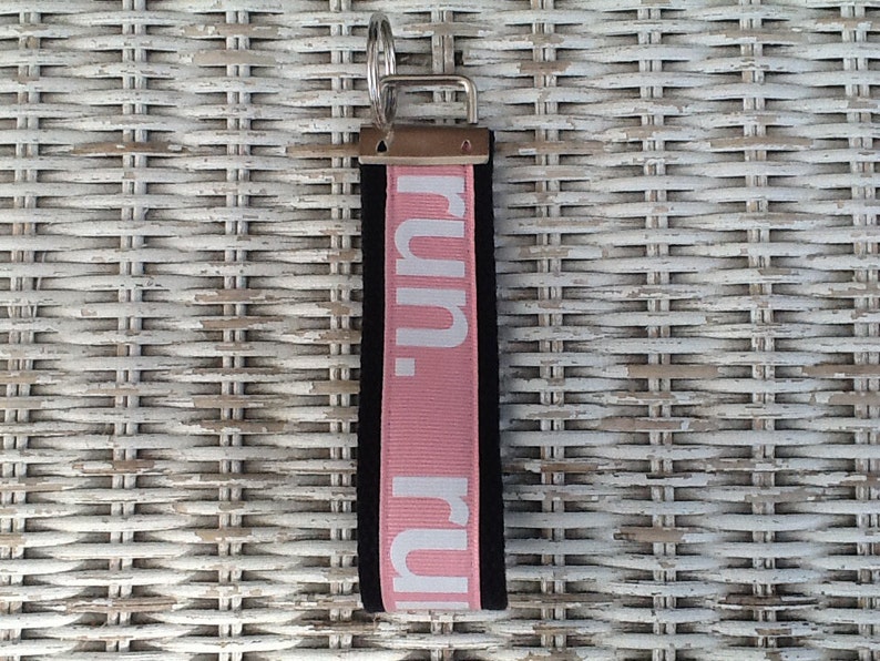 K&ouml;nnte beinhalten: Ein schwarzer und rosa Schl&uuml;sselanh&auml;nger mit dem Wort "run." in wei&szlig;en Buchstaben. Der Schl&uuml;sselanh&auml;nger hat einen silbernen Metallring und eine schwarze R&uuml;ckseite. Der Hintergrund ist eine wei&szlig;e Korboberfl&auml;che.