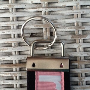 K&ouml;nnte beinhalten: Nahaufnahme eines Schl&uuml;sselbands mit Metallverschluss und Schl&uuml;sselring. Das Schl&uuml;sselband hat einen schwarzen Riemen mit einem rosa Streifen und wei&szlig;em Text. Der Hintergrund ist eine wei&szlig;e Korbgeflechtoberfl&auml;che.