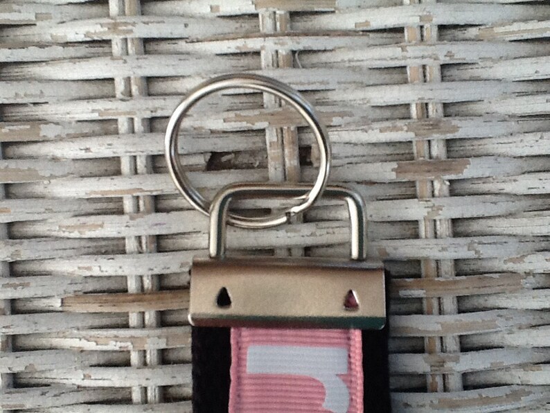 K&ouml;nnte beinhalten: Nahaufnahme eines Schl&uuml;sselbands mit Metallverschluss und Schl&uuml;sselring. Das Schl&uuml;sselband hat einen schwarzen Riemen mit einem rosa Streifen und wei&szlig;em Text. Der Hintergrund ist eine wei&szlig;e Korbgeflechtoberfl&auml;che.
