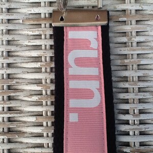 K&ouml;nnte beinhalten: Ein rosa-schwarzer Schl&uuml;sselanh&auml;nger mit dem Wort "run." in Wei&szlig; aufgedruckt. Der Schl&uuml;sselanh&auml;nger hat oben einen Metallverschluss zum Befestigen an Schl&uuml;sseln oder einer Tasche. Der Hintergrund ist eine wei&szlig;e Korboberfl&auml;che.