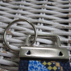 May include: A silver key ring with a ring and a metal clasp. The clasp is attached to a fabric strap with a blue, yellow and white patterned design.