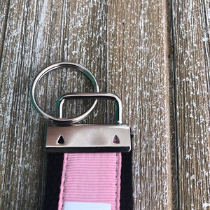K&ouml;nnte beinhalten: Nahaufnahme eines Schl&uuml;sselanh&auml;ngers mit einem silbernen Metallring und einem rechteckigen Metallverschluss. Der Schl&uuml;sselanh&auml;ngerriemen ist schwarz mit einem rosa Streifen und wei&szlig;em Text. Der Hintergrund ist eine verwitterte, hellbraune Holzoberfl&auml;che.