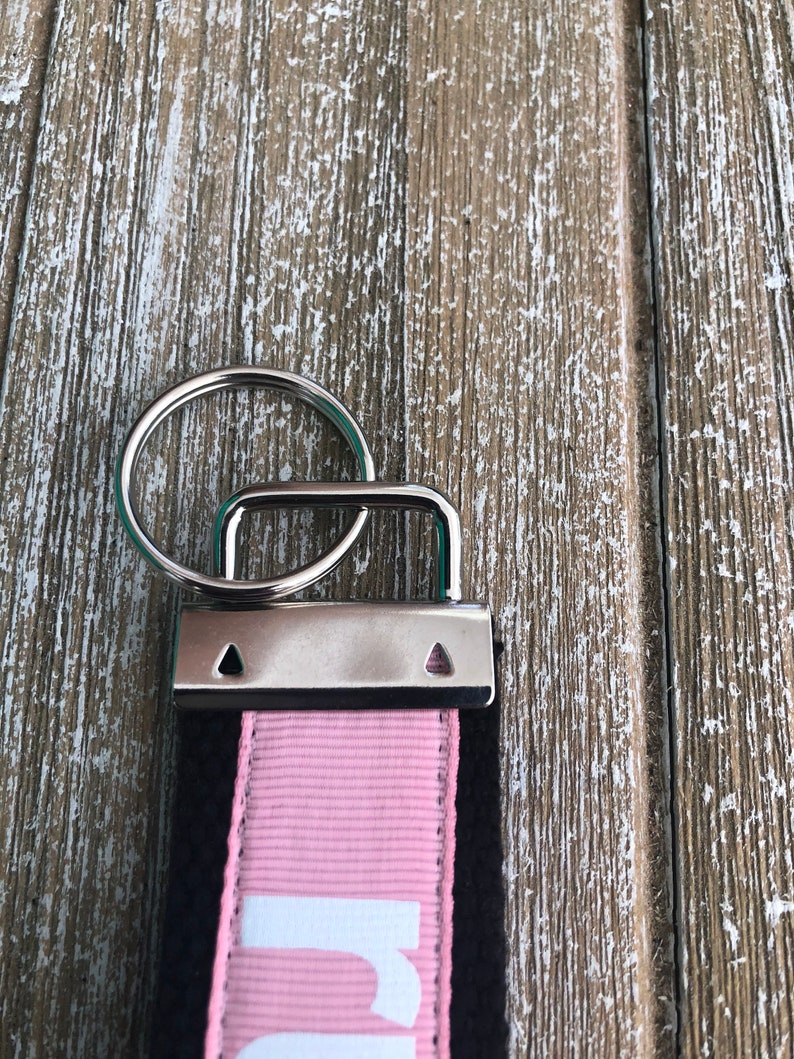 K&ouml;nnte beinhalten: Nahaufnahme eines Schl&uuml;sselanh&auml;ngers mit einem silbernen Metallring und einem rechteckigen Metallverschluss. Der Schl&uuml;sselanh&auml;ngerriemen ist schwarz mit einem rosa Streifen und wei&szlig;em Text. Der Hintergrund ist eine verwitterte, hellbraune Holzoberfl&auml;che.