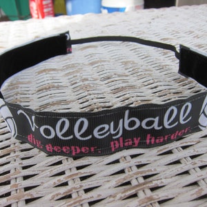 Op de afbeelding: Zwarte hoofdband met het woord "Volleyball" in witte letters, een volleybalafbeelding en de zinnen "dig deeper" en "play harder" in roze. De hoofdband is gemaakt van een lintachtig materiaal.
