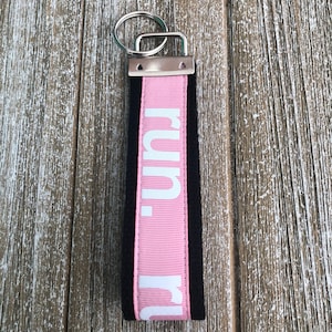 K&ouml;nnte beinhalten: Ein rosa-schwarzes Schl&uuml;sselband mit dem Wort "run" in wei&szlig;en Buchstaben. Das Schl&uuml;sselband besteht aus einem rosa Band mit dem Wort "run" vertikal aufgedruckt, das von schwarzem Stoff umrandet ist. Ein silberner Metallschl&uuml;sselring ist befestigt.