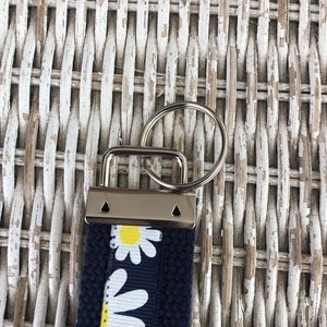 K&ouml;nnte beinhalten: Ein marineblauer Schl&uuml;sselanh&auml;nger mit G&auml;nsebl&uuml;mchenmuster. Der Schl&uuml;sselanh&auml;nger hat einen silberfarbenen Metallverschluss und einen Spaltring zum Anbringen von Schl&uuml;sseln. Das G&auml;nsebl&uuml;mchenmuster hat wei&szlig;e Bl&uuml;tenbl&auml;tter und gelbe Zentren.