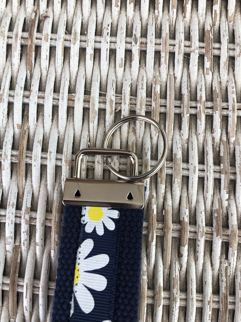 K&ouml;nnte beinhalten: Ein marineblauer Schl&uuml;sselanh&auml;nger mit G&auml;nsebl&uuml;mchenmuster. Der Schl&uuml;sselanh&auml;nger hat einen silberfarbenen Metallverschluss und einen Spaltring zum Anbringen von Schl&uuml;sseln. Das G&auml;nsebl&uuml;mchenmuster hat wei&szlig;e Bl&uuml;tenbl&auml;tter und gelbe Zentren.
