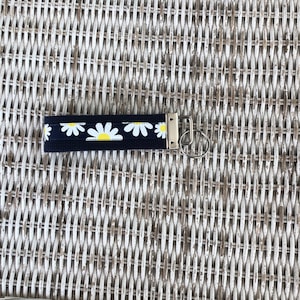 K&ouml;nnte beinhalten: Ein marineblaues Schl&uuml;sselband mit einem Muster aus wei&szlig;en G&auml;nsebl&uuml;mchen und gelben Zentren. Es hat einen silberfarbenen Metallverschluss und Schl&uuml;sselring. Der Hintergrund ist eine geflochtene Korbwarenoberfl&auml;che.