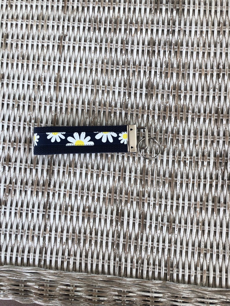 K&ouml;nnte beinhalten: Ein marineblaues Schl&uuml;sselband mit einem Muster aus wei&szlig;en G&auml;nsebl&uuml;mchen und gelben Zentren. Es hat einen silberfarbenen Metallverschluss und Schl&uuml;sselring. Der Hintergrund ist eine geflochtene Korbwarenoberfl&auml;che.