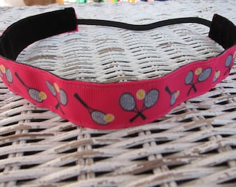 May include: A pink headband with a black elastic band and a white and grey tennis racket design. The headband has yellow tennis balls on it.