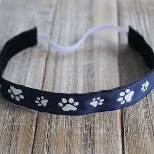 May include: A navy blue headband with white paw print designs. The headband is made of elastic material and is designed to fit comfortably on the head.
